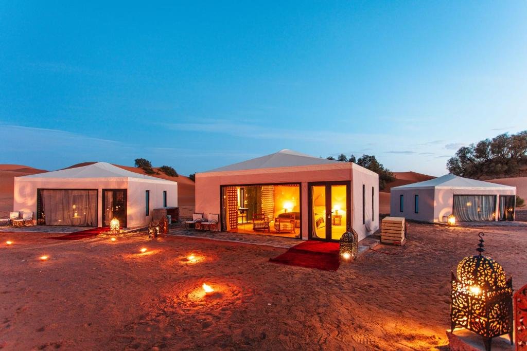 Luxury desert camp lit up at night in the Moroccan Sahara, with glowing tents and traditional lanterns under a twilight sky