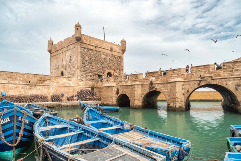 Blue fishing boats docked at the port of Essaouira, Morocco with Skala fortress and stone arch bridge in the background