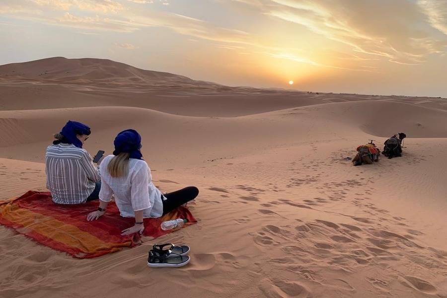 Two travelers sitting on a carpet in the Sahara Desert watching the sunset over the dunes near Merzouga, Morocco