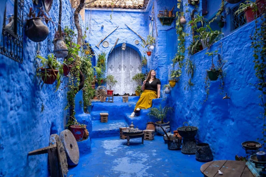 Woman in a yellow dress sitting in a bright blue alleyway in Chefchaouen, Morocco surrounded by traditional decor and flower pots